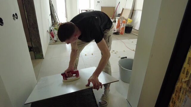 A worker lays large tiles in a commercial space under construction. As the work continues, the space fills with tools and materials. Signs of renovation and preparation are visible throughout the spac