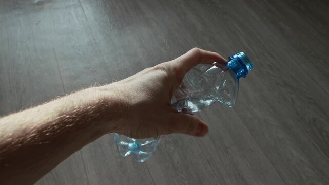 Male hand firmly squeezes empty transparent plastic bottle, creating crinkled effect and reducing volume. Scene highlights consumer waste, importance of recycling, and environmental responsibility.