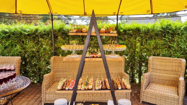 Outdoor catering display features diverse selection of gourmet finger food. Multi-tiered stand presents sweet desserts, savory canap s, and small bites. Wicker furniture and lush green hedge complete 