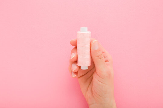 Young adult woman hand holding and showing new pink thread spool on light pink table background. Pastel color. Closeup. Top down view.