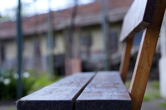 Close-up wooden bench edge with shallow depth of field, blurred garden and building background, calm outdoor atmosphere in daylight 2026