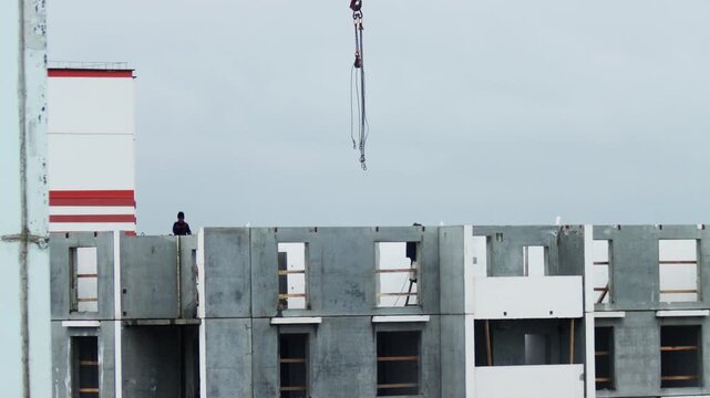 Urban construction site view of residential building under development, workers on top of concrete panel structure, crane hook for lifting operations, grey sky backdrop