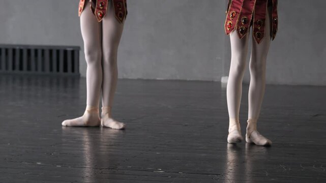 Two young ballerinas in red costumes dance on stage. Feet positioned en pointe on dark wooden floor. Gray wall and radiator visible behind. Motion suggests rehearsal or performance
