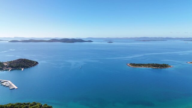 Drone sliding parallel with the horizon over the clear blue Adriatic, passing small islands before revealing Drage marina with white piers and boats on a sunny day.