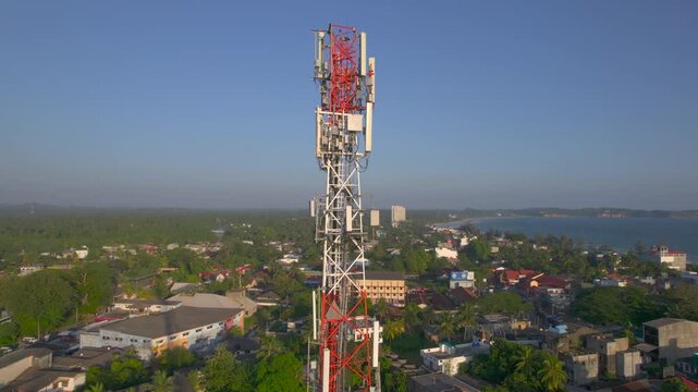 Aerial video of telecom cell tower rising above tropical coastal town with palm trees seaside buildings and calm bay communication infrastructure and shoreline landscape in daylight close view