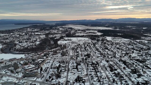 Picturesque aerial perspective flying over a plattsburgh, new york covered in fresh snow during a beautiful winter sunrise, revealing a tranquil and cold urban landscape with a lake in the background