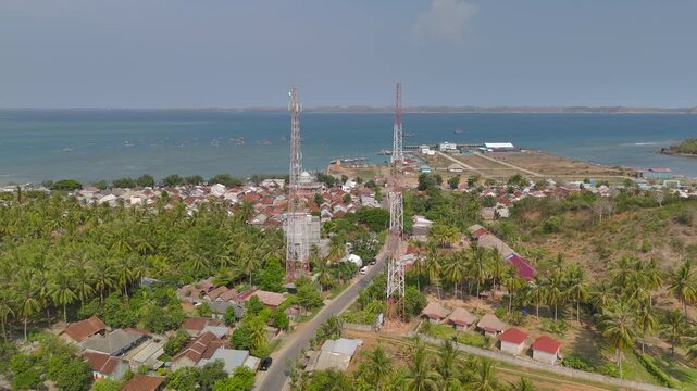 coastal village Aerial video of twin telecom towers above tropical with palm groves rooftops winding road and calm bay showing shoreline infrastructure and regional connectivity in daylight