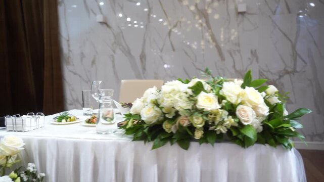 Wedding table decor with white flowers, blank card stand on white tablecloth, fabric drapery background, floral arrangement front view, with copy space. Concept wedding, ceremony, invitation, luxury