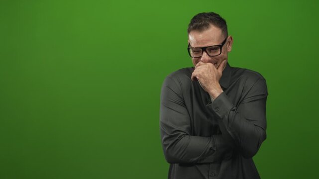 Man with hand covering mouth and crossed arms, stifling laugh while looking down against green screen studio; amusement.