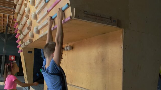 Boy hangs on horizontal bar at bottom of climbing wall. Young athlete warms up before starting training in gym slow motion