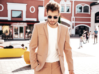 Handsome confident stylish hipster lambersexual model.Sexy modern man dressed in elegant beige suit. Fashion male posing in the street background in Europe city at sunset. In sunglasses © halayalex