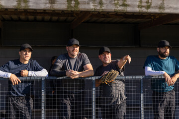 Four men wearing caps standing by wire fence in dugout, pointing toward field with glove © wavebreak3