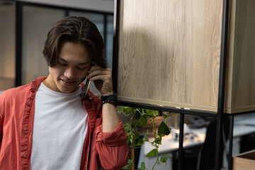 Adult Asian man wearing red shirt, standing on phone call in coworking space, holding smartphone © wavebreak3