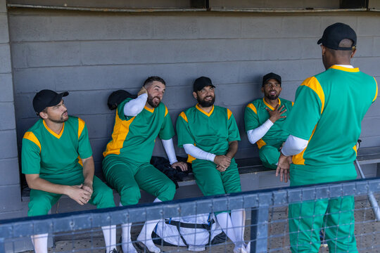 Male baseball teammates sitting on bench in dugout wearing green-yellow uniforms near duffel bag