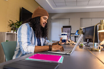 Male worker in beanie typing on laptop while holding blue-white coffee cup at desk near notebooks © wavebreak3