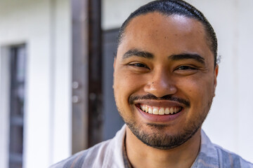 Asian man smiling and facing camera on porch in collared striped shirt with doorframe and window © wavebreak3