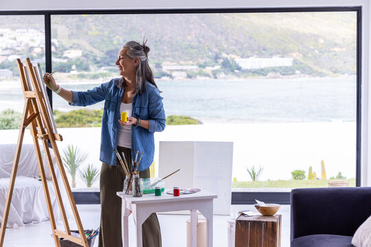 Senior woman painting at easel in coastal home wearing denim with yellow paint jar, copy space