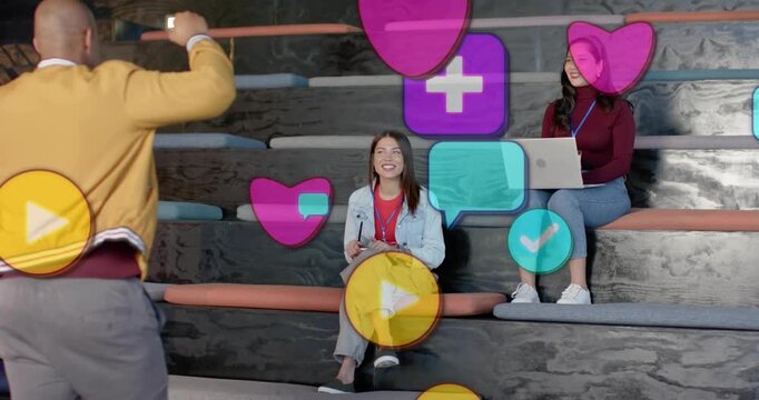 Man presenting tech app and gesturing, causing AR icons with pink hearts engaging seated women