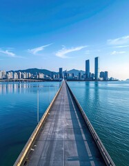 Fototapeta premium A long concrete bridge stretches across a calm bay towards a modern cityscape with towering skyscrapers and distant mountains under a clear blue sky.