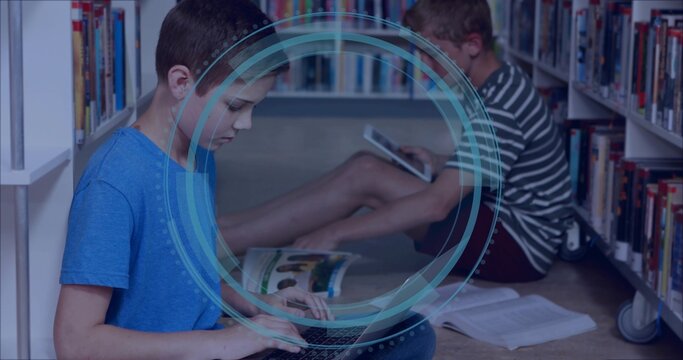 Typing child wearing blue T-shirt, classmate using tablet in library aisle with laptop and books