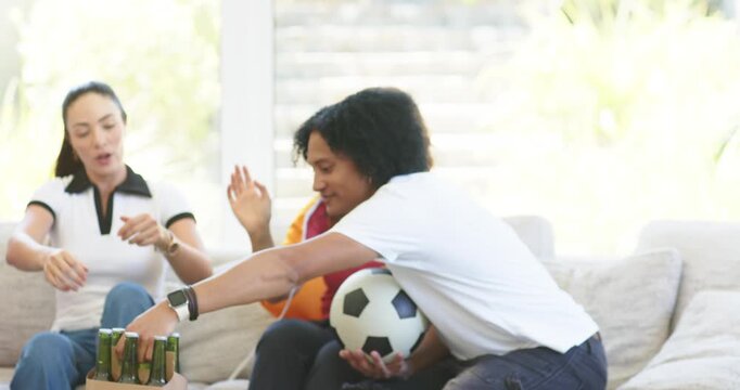 Group of four friends watching sports on couch, passing bottles from carrier, holding soccer ball