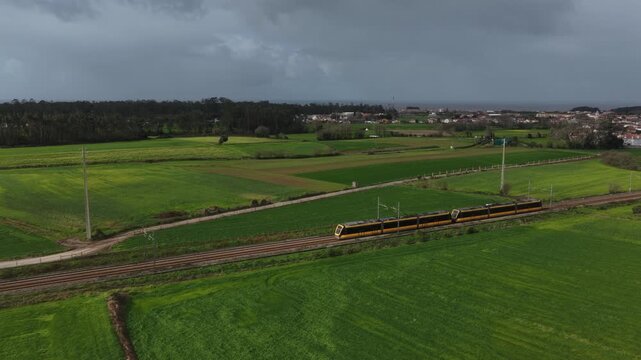 Aerial view of a vibrant yellow train slicing through lush green fields under a moody sky, Vila do Conde, Portugal.