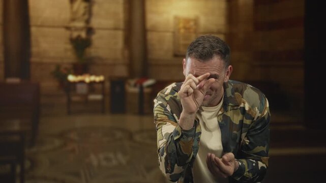Man soldier in camouflage jacket holding clasped hands and pleading gesture inside a church building; contrition sorrow.