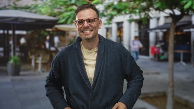 Man in robe smiling with hands on hips amid street cafe tables and trees, wearing glasses and t shirt, standing on a paved pedestrian plaza; confidence ease.