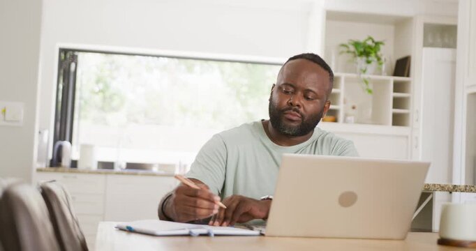 African American man hearing sound glancing writing notes working in kitchen laptop notebook pencil