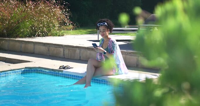 Adult female holding device at pool edge with sunglasses, tapping screen and relaxing, feet in pool