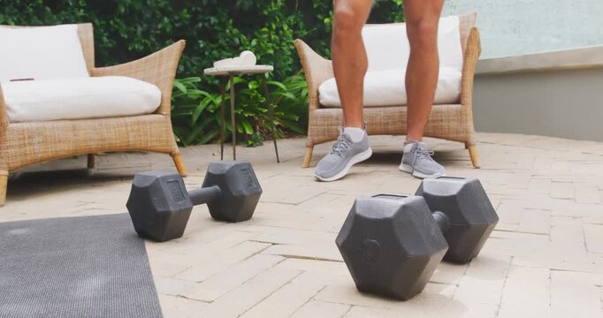 Male in shorts shifting, pushing up from chair on paved patio, grasping dumbbells, preparing set
