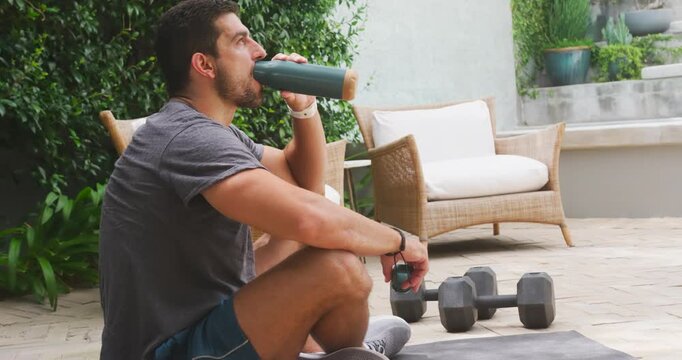 Man drinking from bottle post workout, inspecting cap while resting on patio mat near dumbbells