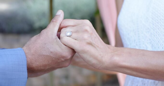 Couple after ring exchange, man in blue shirt steadying woman's hand presenting halo ring outdoors