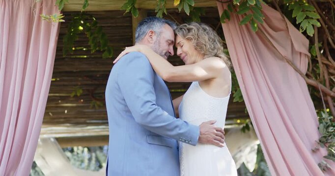 Newlywed couple in formal wear holding forehead touch under pergola with pink drapes, then smiling