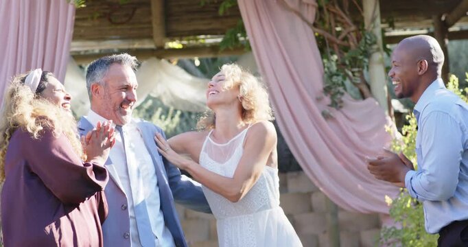 Couple in formal wear with diverse guests after applause at pergola, bride twirling, groom laughing