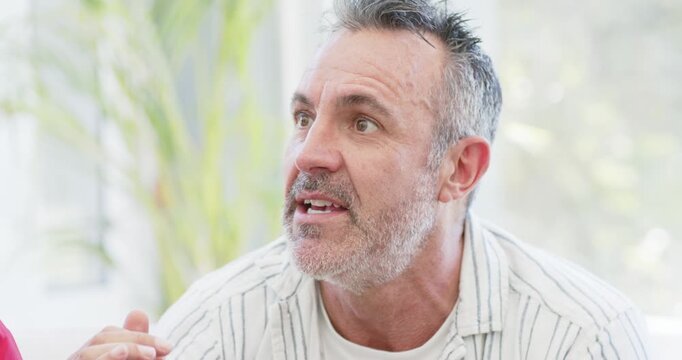 Middle-aged man speaking, pinching fingers and reacting to off-camera comment near sunroom plant