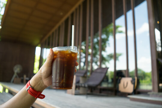 Hand hold ice coffee glass.Drink black or americano coffee at the park of cafe restaurant with glass building and sunlight background
