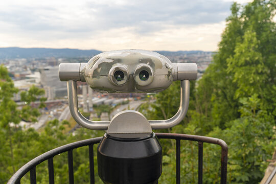 View of a silver coin operated telescope with two eyepieces fixed on a metal railing, overlooking the distant city skyline and lush green trees, Oslo, Norway.