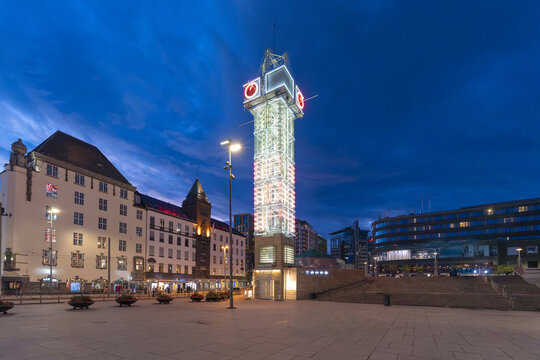 View of a tower gleaming with lights against the deep blue twilight sky, surrounded by modern buildings, offering a striking contrast of old and new, Oslo, Norway.