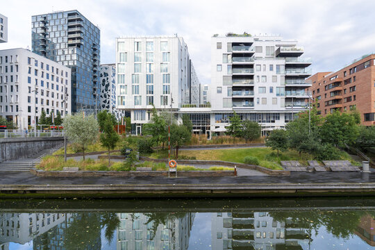 View of modern buildings reflected in the canal's shimmering surface, framed by lush greenery and contemporary architecture, Oslo, Norway.