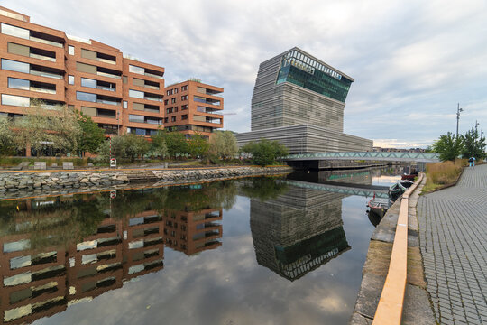 View of modern architecture reflected in the serene waters of a canal under a cloudy sky, where brick buildings meet the unique design of Munch museum, Oslo, Norway.
