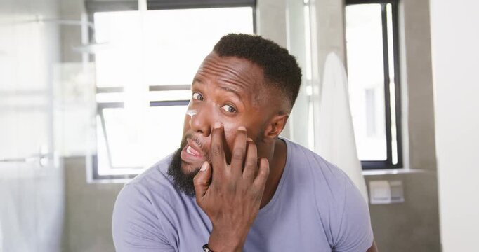 African American man applying and blending facial cream at bathroom sink wearing lavender t-shirt