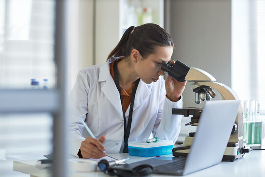 Young adult Caucasian woman scientist examining sample through microscope and taking notes in laboratory. Ideal for medical research, biotech innovation, diagnostics marketing