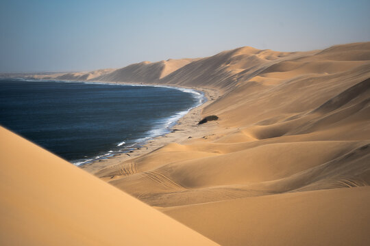 View of rolling golden sand dunes meet the deep blue sea under a clear sky, creating a striking contrast in Anichab, Erongo, Namibia.
