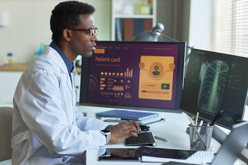 Fototapeta premium Black male doctor reviewing digital patient records and chest scan on desktop monitors, supporting telemedicine workflow. Ideal for healthcare technology, diagnostics, medical data themes
