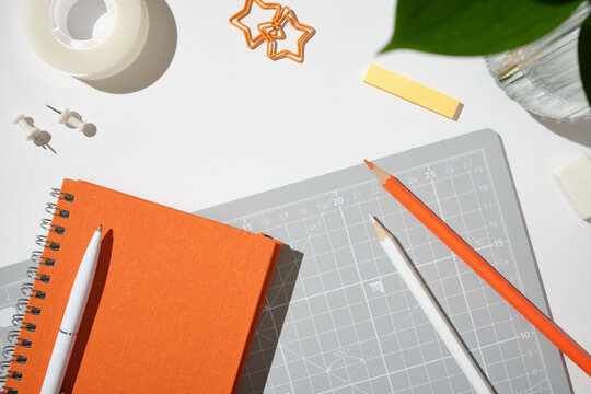 Top view of a closed orange notebook, white pen, cutting mat, and pencils on a white background. Copyspace
