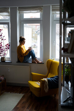 Woman sitting on a windowsill at home using a smartphone and drinking tea