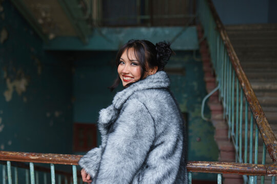 Smiling woman in fur coat near old staircase with shabby blue walls