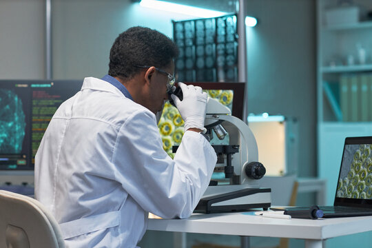 African American male scientist examining specimen through microscope in laboratory, analyzing medical data on screens. Useful for healthcare research, diagnostics, biotech marketing