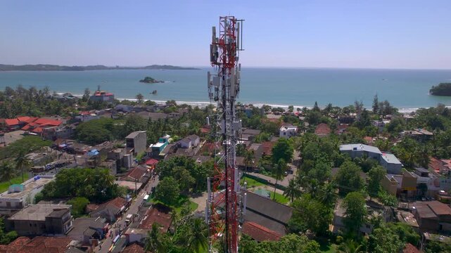 Aerial video of telecom cell tower above tropical seaside town with beach palms rooftops calm bay and small rocky islets showing coastal communication infrastructure in daylight life in a coastal